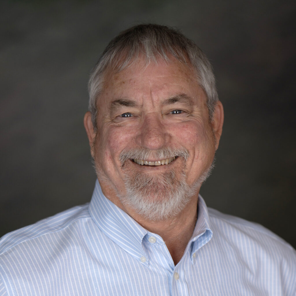 A smiling older man with a beard, wearing a light blue striped shirt, against a blurred dark background.